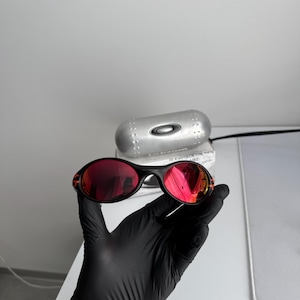 May include: Black sunglasses with red lenses. The sunglasses are held by a gloved hand. The frame is black, and the lenses reflect the surroundings. A silver case is visible in the background, suggesting a protective storage solution.