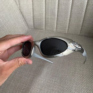May include: Silver-framed sunglasses with dark lenses. The sunglasses have a sleek, modern design with a curved frame and temples. The lenses are oval-shaped and appear to be tinted. The sunglasses are held by a hand.