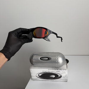 May include: Black sunglasses with red-tinted lenses. The sunglasses are held above a silver case and a white box with the text "SCIENCE AND ART". The case has a metallic finish and the box has a logo.
