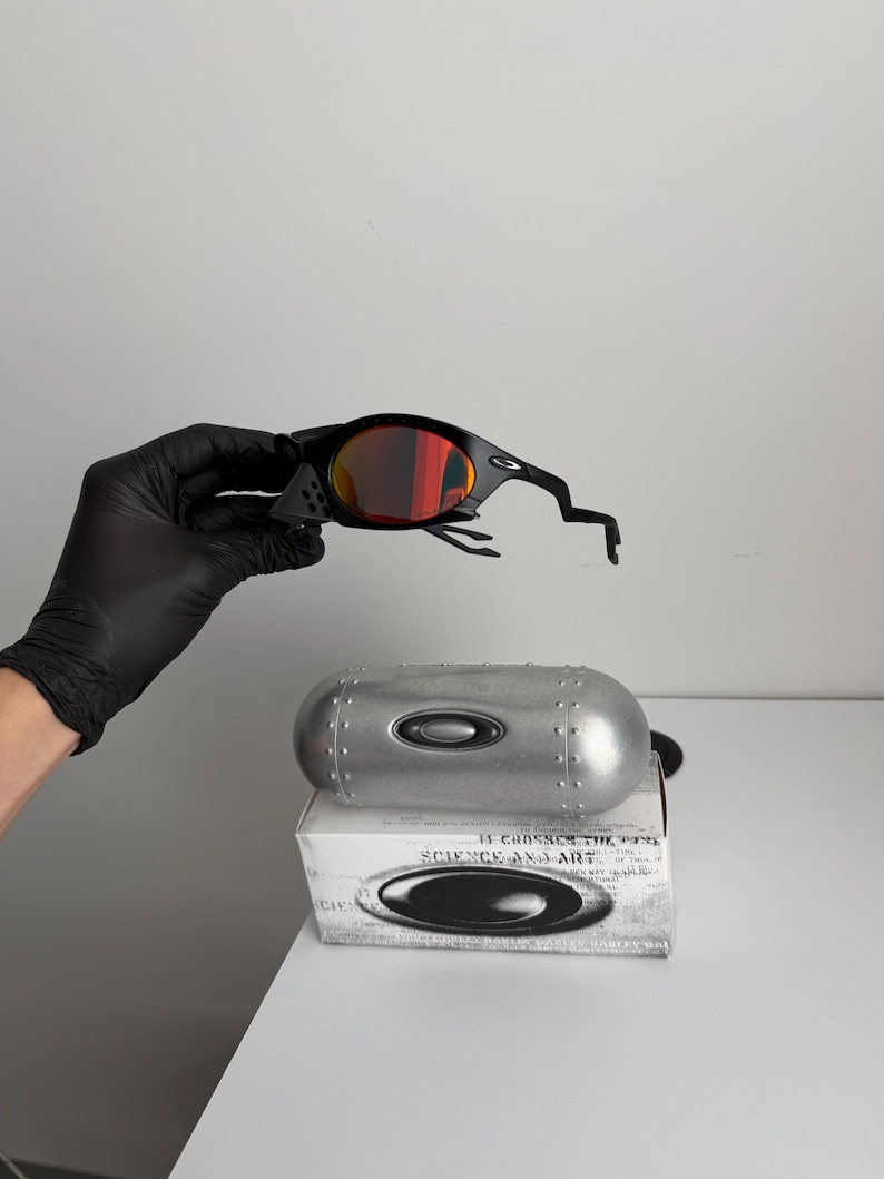 May include: Black sunglasses with red-tinted lenses. The sunglasses are held above a silver case and a white box with the text "SCIENCE AND ART". The case has a metallic finish and the box has a logo.