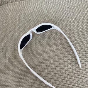 Puede incluir: Gafas de sol blancas con lentes negras. Las gafas tienen un dise&ntilde;o elegante y moderno con una montura curvada y una forma aerodin&aacute;mica. Las lentes son oscuras, proporcionando contraste con la montura blanca.