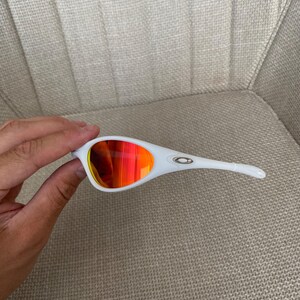 Puede incluir: Gafas de sol blancas con lentes rojo-naranja. Las gafas de sol tienen un dise&ntilde;o elegante y moderno con montura y patillas blancas. Las lentes son de un vibrante color rojo-naranja, proporcionando un contraste llamativo con la montura blanca. El logotipo de Oakley es visible en la patilla.
