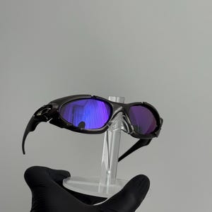 May include: Black sunglasses with purple-tinted lenses are displayed on a clear stand. The sunglasses have a sleek, sporty design with curved arms and a dark frame. The lenses reflect a vibrant purple hue.