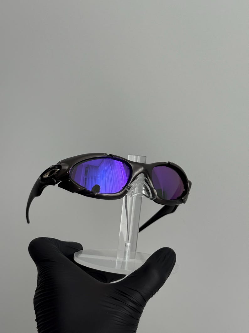 May include: Black sunglasses with purple-tinted lenses are displayed on a clear stand. The sunglasses have a sleek, sporty design with curved arms and a dark frame. The lenses reflect a vibrant purple hue.