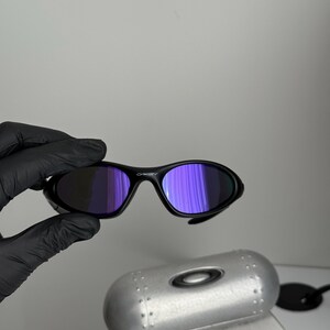 May include: Black sunglasses with purple-tinted lenses. The sunglasses have a sleek, curved design. The image also shows a silver case and a box with the text "SCIENCE AND ART".