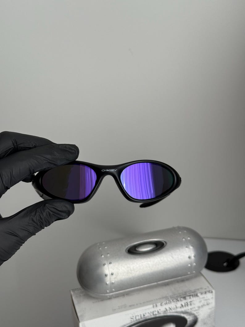 May include: Black sunglasses with purple-tinted lenses. The sunglasses have a sleek, curved design. The image also shows a silver case and a box with the text "SCIENCE AND ART".