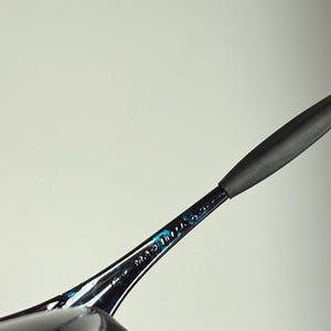 Puede incluir: Primer plano de una montura de gafas negra y azul. Las palabras "MADE IN U.S.A." son visibles en la varilla. La montura tiene un dise&ntilde;o elegante y moderno con una patilla de goma negra.