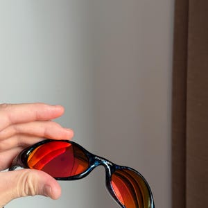 Puede incluir: Gafas de sol negras y azules con lentes espejadas rojas. Las gafas de sol tienen un dise&ntilde;o curvo y el texto "BRAIN DEAD" est&aacute; impreso en la montura. Las lentes reflejan el entorno.