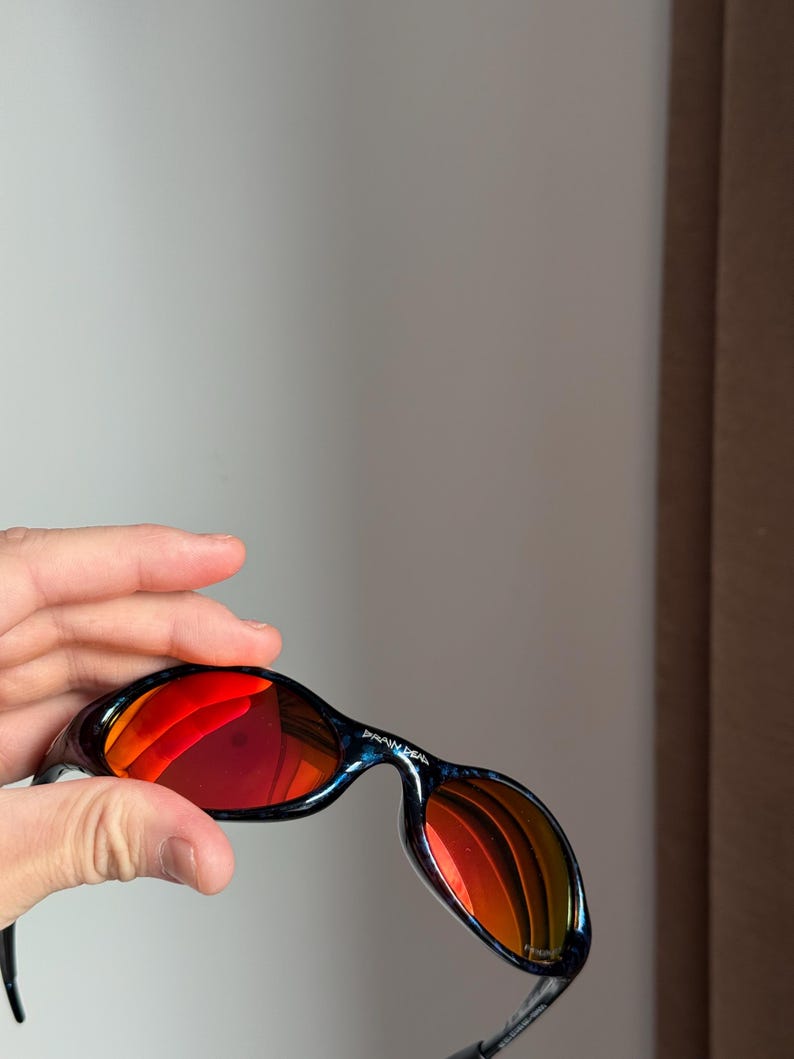 Puede incluir: Gafas de sol negras y azules con lentes espejadas rojas. Las gafas de sol tienen un dise&ntilde;o curvo y el texto "BRAIN DEAD" est&aacute; impreso en la montura. Las lentes reflejan el entorno.