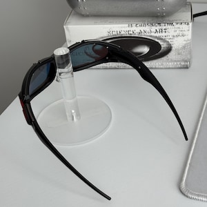 May include: Black sunglasses with blue lenses are displayed on a clear stand. The sunglasses have a red accent on the frame. A silver case and a box with the text "SCIENCE AND ART" are in the background.