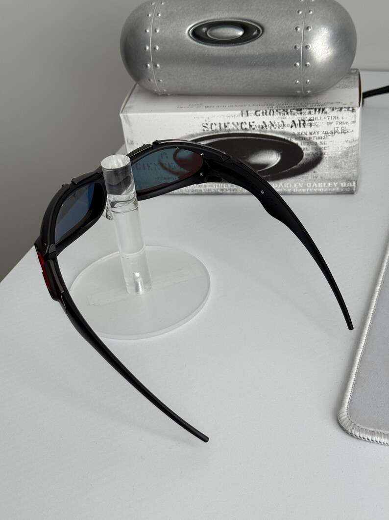 May include: Black sunglasses with blue lenses are displayed on a clear stand. The sunglasses have a red accent on the frame. A silver case and a box with the text "SCIENCE AND ART" are in the background.