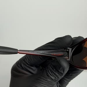 May include: Close-up of sunglasses with a brown and black frame. The word "Jacket" is printed on the arm of the sunglasses. The sunglasses are held by a person wearing black gloves.