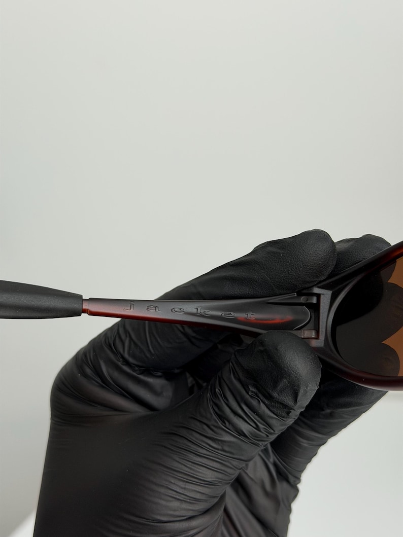 May include: Close-up of sunglasses with a brown and black frame. The word "Jacket" is printed on the arm of the sunglasses. The sunglasses are held by a person wearing black gloves.