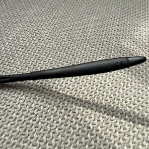 May include: A black eyeglass arm with a rounded, tapered design. The arm has a matte finish and the letters "CE" are visible near the tip. The background is a textured, light grey fabric.