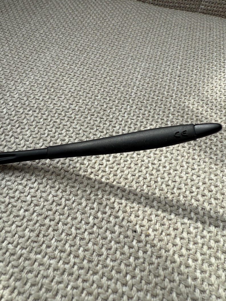 May include: A black eyeglass arm with a rounded, tapered design. The arm has a matte finish and the letters "CE" are visible near the tip. The background is a textured, light grey fabric.