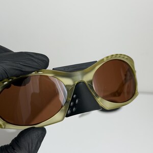 May include: Olive green sunglasses with brown lenses. The sunglasses have a unique, wraparound design with a black nose bridge and temples. The lenses are a warm brown colour, and the frames have a textured appearance.