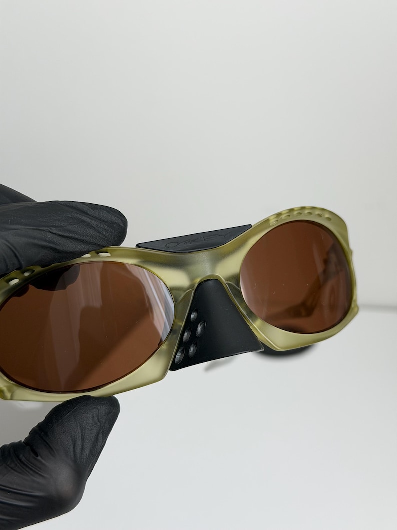 May include: Olive green sunglasses with brown lenses. The sunglasses have a unique, wraparound design with a black nose bridge and temples. The lenses are a warm brown colour, and the frames have a textured appearance.