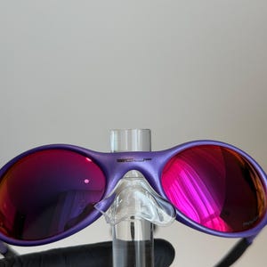 May include: Purple sunglasses with reflective pink lenses. The sunglasses have a matte finish and a clear nose bridge. The temples are black. The brand name is visible on the top of the frame.