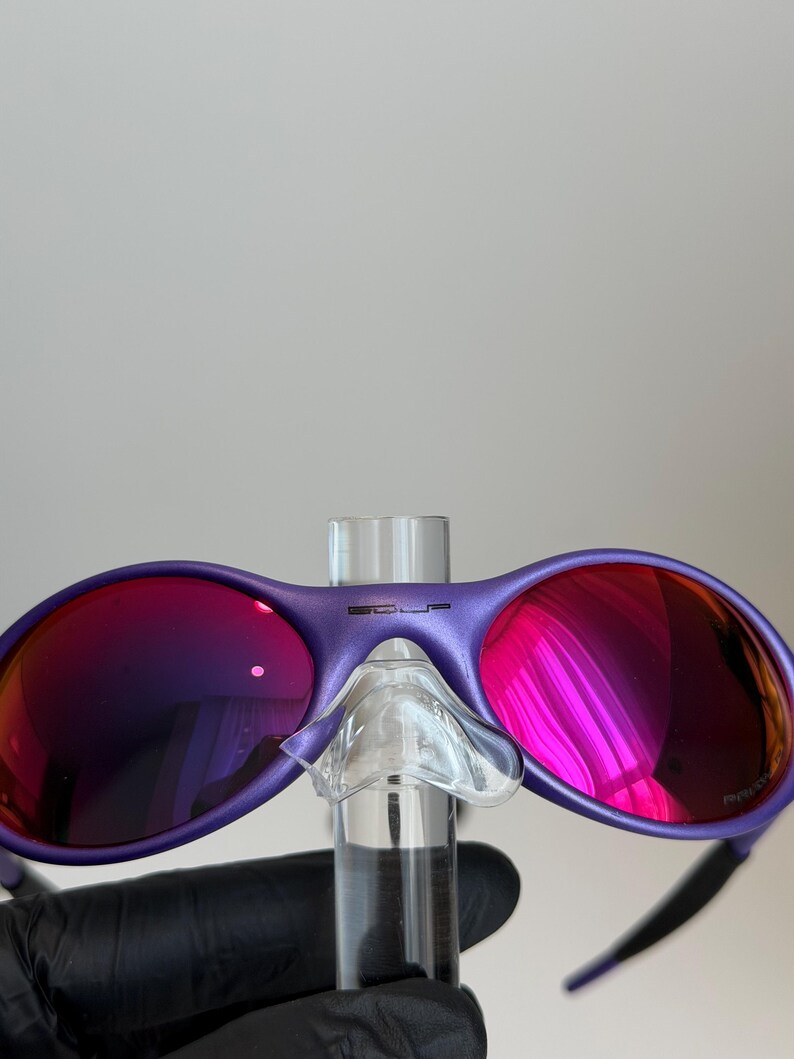May include: Purple sunglasses with reflective pink lenses. The sunglasses have a matte finish and a clear nose bridge. The temples are black. The brand name is visible on the top of the frame.