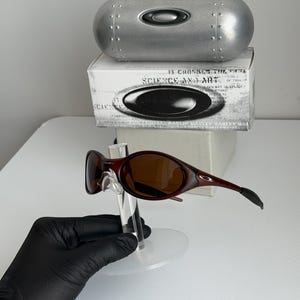 May include: Red-brown sunglasses with brown lenses are displayed on a clear stand. A silver case and a white box with black text and a logo are in the background. The sunglasses have a sleek, modern design.