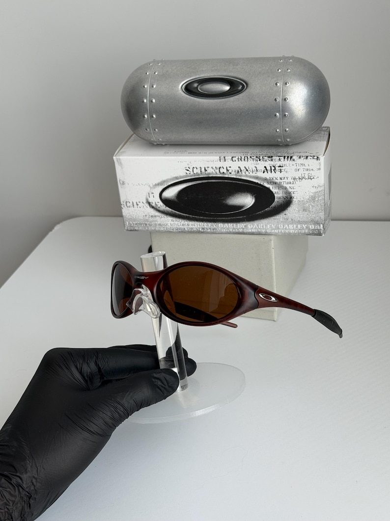 May include: Red-brown sunglasses with brown lenses are displayed on a clear stand. A silver case and a white box with black text and a logo are in the background. The sunglasses have a sleek, modern design.