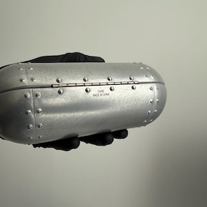 May include: A silver-colored, riveted case with a hinged lid. The case has a rounded, cylindrical shape and is held by a gloved hand. The text "CASE MADE IN CHINA" is visible on the case.