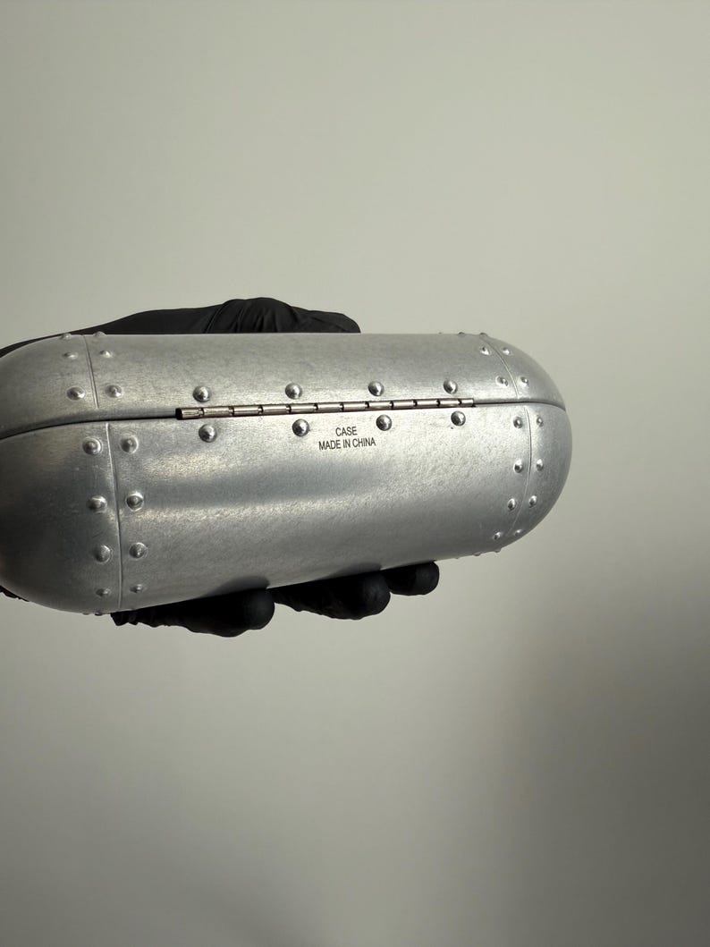 May include: A silver-colored, riveted case with a hinged lid. The case has a rounded, cylindrical shape and is held by a gloved hand. The text "CASE MADE IN CHINA" is visible on the case.