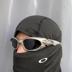 May include: Silver-framed sunglasses with dark lenses. The sunglasses are worn over a black balaclava with a white logo. The balaclava covers the head and neck, leaving only the eyes visible.