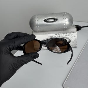 Puede incluir: Gafas de sol negras con lentes marrones. Las gafas de sol est&aacute;n sobre una caja blanca con el texto "SCIENCE AND ART". Una funda plateada con el logotipo de Oakley est&aacute; detr&aacute;s de las gafas.