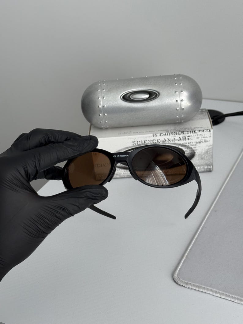 Puede incluir: Gafas de sol negras con lentes marrones. Las gafas de sol est&aacute;n sobre una caja blanca con el texto "SCIENCE AND ART". Una funda plateada con el logotipo de Oakley est&aacute; detr&aacute;s de las gafas.