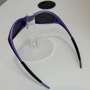 May include: Purple sunglasses with black lenses and black earpieces are displayed on a clear stand. The sunglasses have a sleek, modern design with a curved frame. The earpieces have a textured grip for comfort and security.