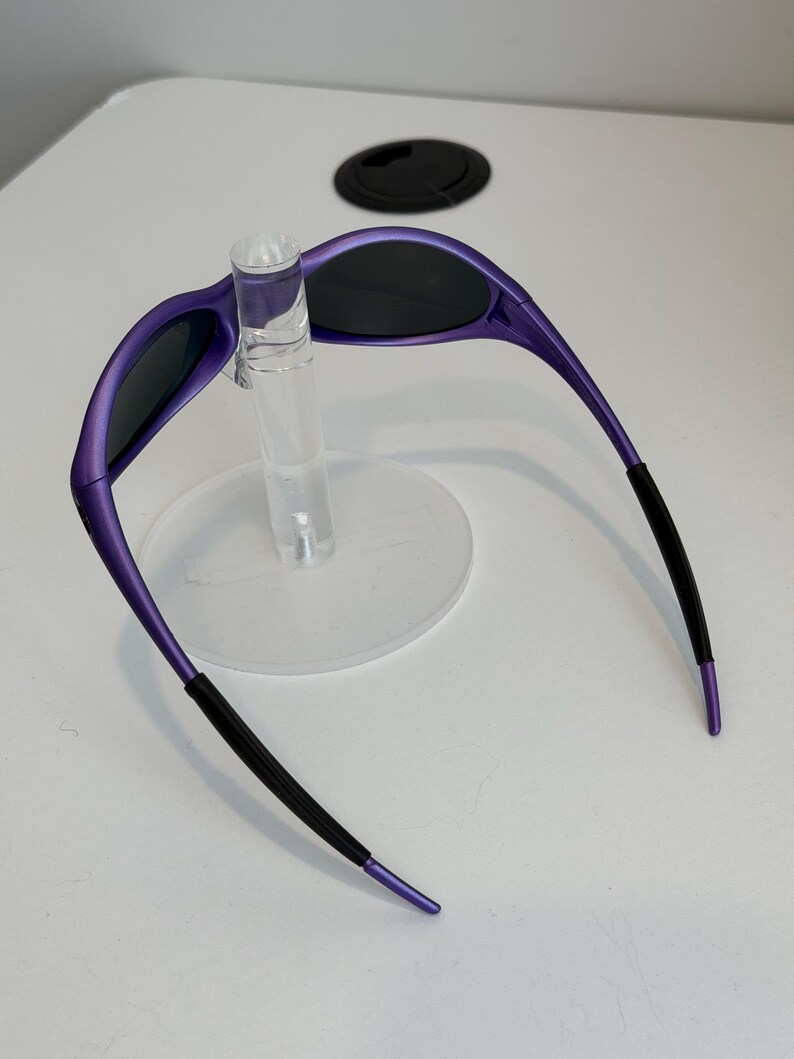 May include: Purple sunglasses with black lenses and black earpieces are displayed on a clear stand. The sunglasses have a sleek, modern design with a curved frame. The earpieces have a textured grip for comfort and security.