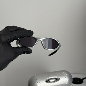 Puede incluir: Gafas de sol con montura plateada y lentes oscuras. Las gafas de sol est&aacute;n sujetas por una mano enguantada. Debajo de las gafas de sol hay un estuche plateado con el logotipo de Oakley. El estuche est&aacute; encima de una caja con el texto "SCIENCE AND ART."