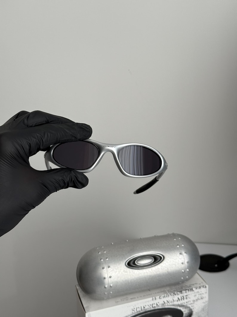 Puede incluir: Gafas de sol con montura plateada y lentes oscuras. Las gafas de sol est&aacute;n sujetas por una mano enguantada. Debajo de las gafas de sol hay un estuche plateado con el logotipo de Oakley. El estuche est&aacute; encima de una caja con el texto "SCIENCE AND ART."