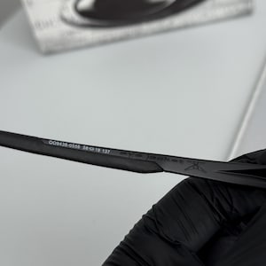Puede incluir: Primer plano de gafas de sol negras con el texto "eye jacket" y los n&uacute;meros "009438-0558 56 19 137" impresos en la patilla. Las gafas de sol est&aacute;n sobre un fondo blanco.