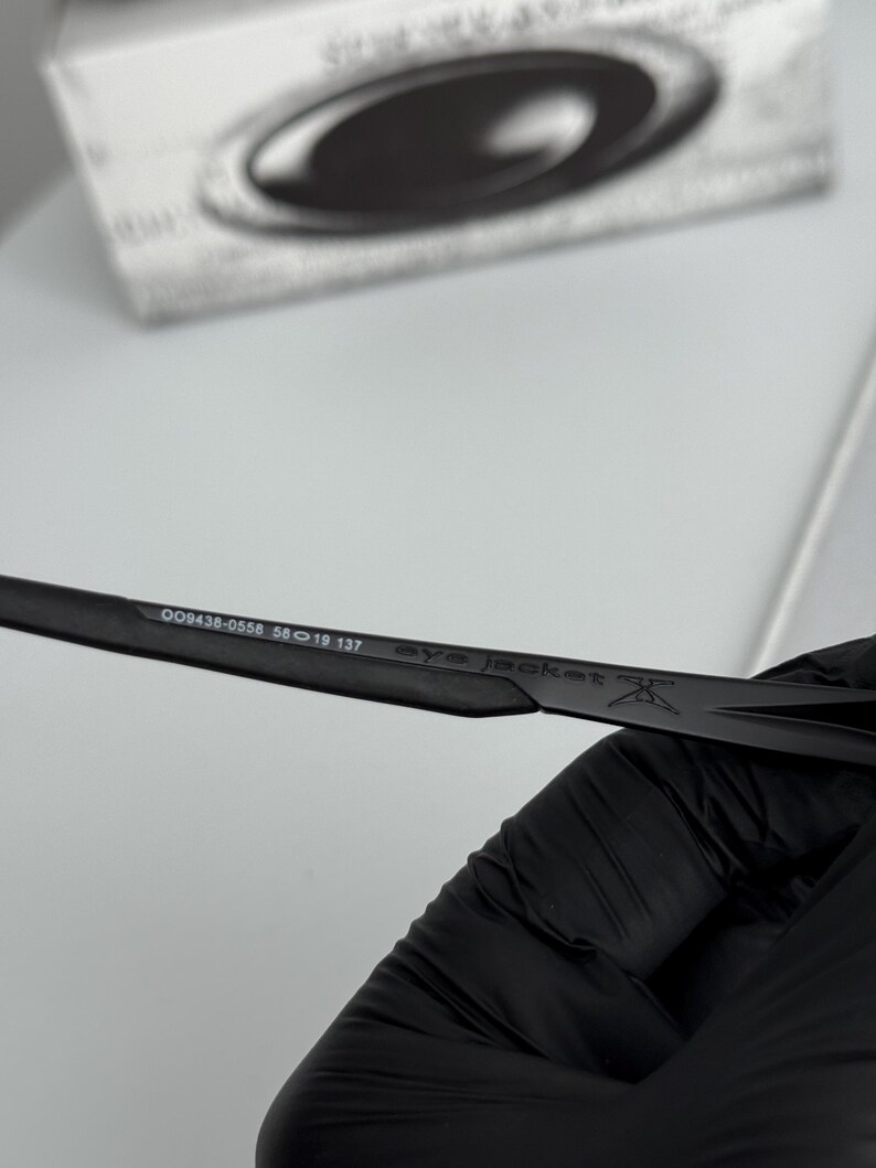 Puede incluir: Primer plano de gafas de sol negras con el texto "eye jacket" y los n&uacute;meros "009438-0558 56 19 137" impresos en la patilla. Las gafas de sol est&aacute;n sobre un fondo blanco.