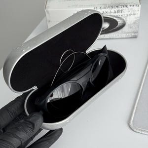 K&ouml;nnte beinhalten: Schwarze Sonnenbrille in einem silbernen Etui. Das Etui ist ge&ouml;ffnet und zeigt die Sonnenbrille mit schwarzem Innenraum. Im Hintergrund ist eine wei&szlig;e Box mit Text zu sehen. Modernes Design.