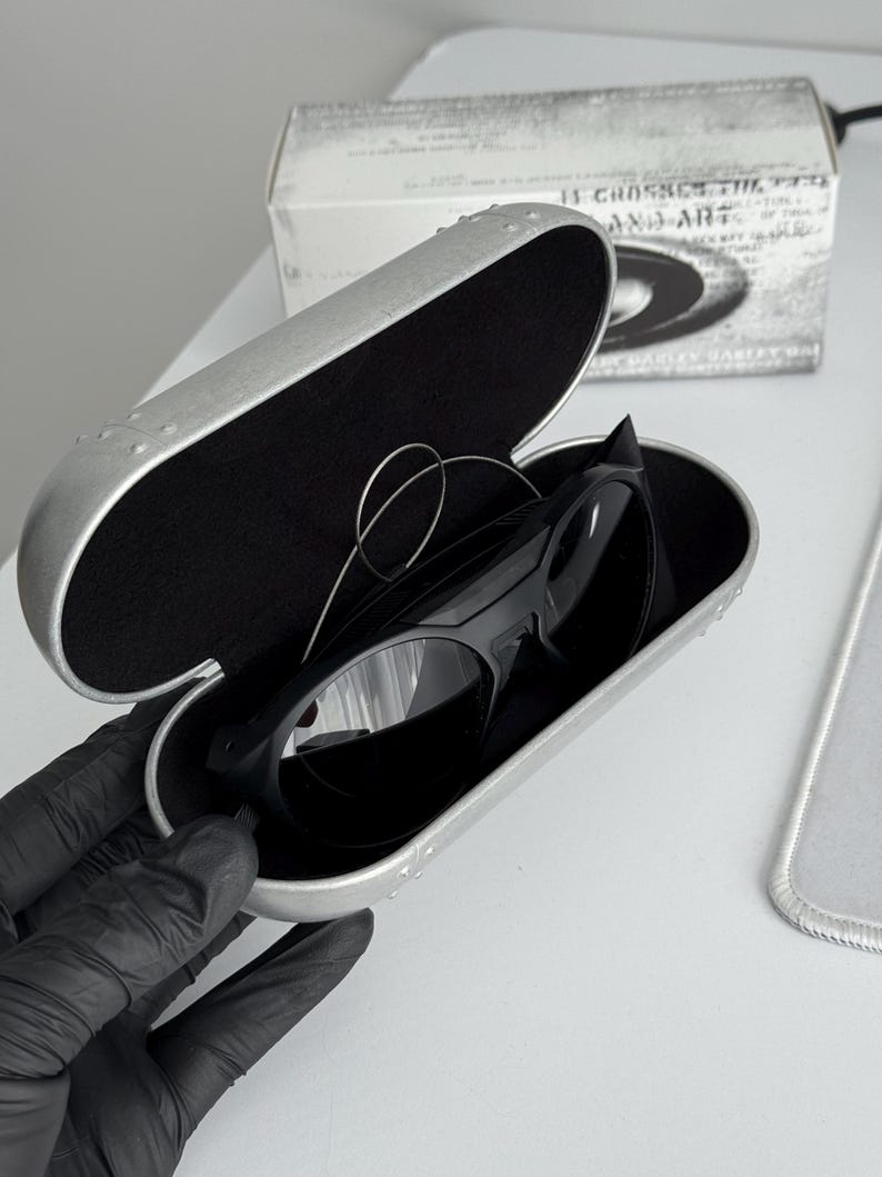 K&ouml;nnte beinhalten: Schwarze Sonnenbrille in einem silbernen Etui. Das Etui ist ge&ouml;ffnet und zeigt die Sonnenbrille mit schwarzem Innenraum. Im Hintergrund ist eine wei&szlig;e Box mit Text zu sehen. Modernes Design.