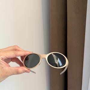 May include: Beige-framed sunglasses with dark lenses. The sunglasses have a rounded shape and are held by a hand. The background includes a white wall and brown curtains.