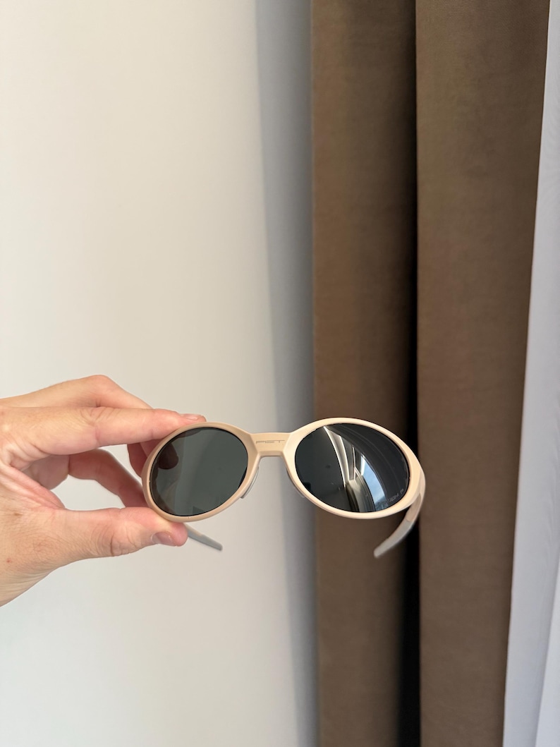 May include: Beige-framed sunglasses with dark lenses. The sunglasses have a rounded shape and are held by a hand. The background includes a white wall and brown curtains.