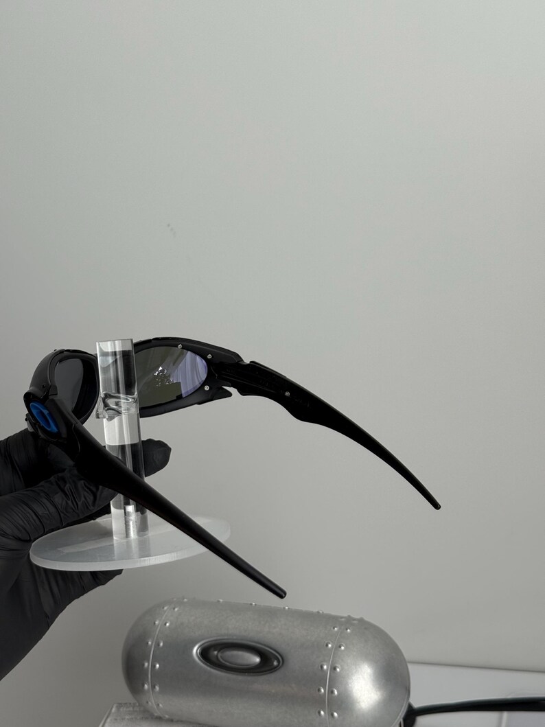 May include: Black sunglasses with a silver mirrored lens and a matching case. The sunglasses have a sleek, modern design with a curved frame and arms. The case is cylindrical with a metallic finish.