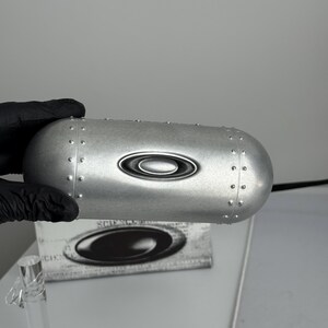 May include: A silver-colored, riveted eyewear case with the Oakley logo. The case has a rounded, oblong shape and a metallic finish. The Oakley logo is centered on the case. The case is held by a gloved hand.