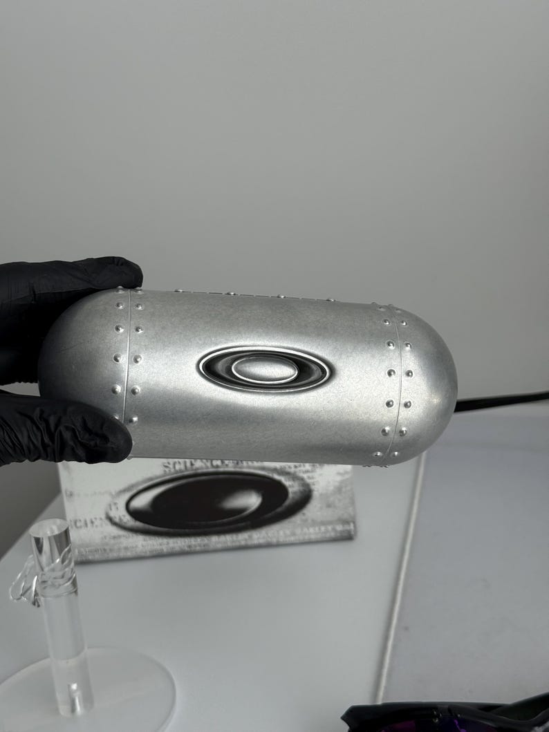 May include: A silver-colored, riveted eyewear case with the Oakley logo. The case has a rounded, oblong shape and a metallic finish. The Oakley logo is centered on the case. The case is held by a gloved hand.