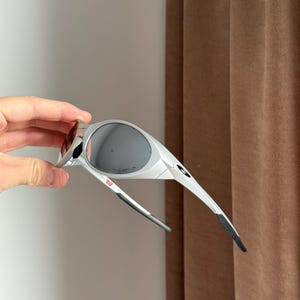 May include: White and grey sports sunglasses with dark lenses. The sunglasses have a sleek design with a small red accent on the frame. The arms are thin and taper to a black tip.