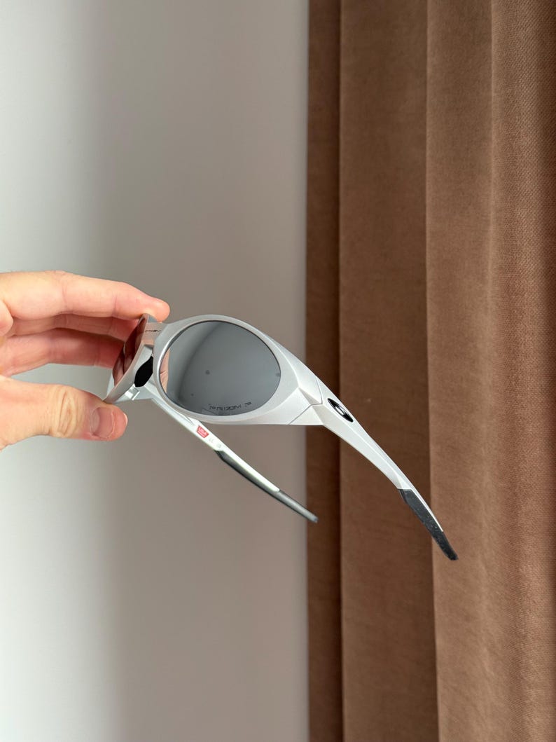 May include: White and grey sports sunglasses with dark lenses. The sunglasses have a sleek design with a small red accent on the frame. The arms are thin and taper to a black tip.