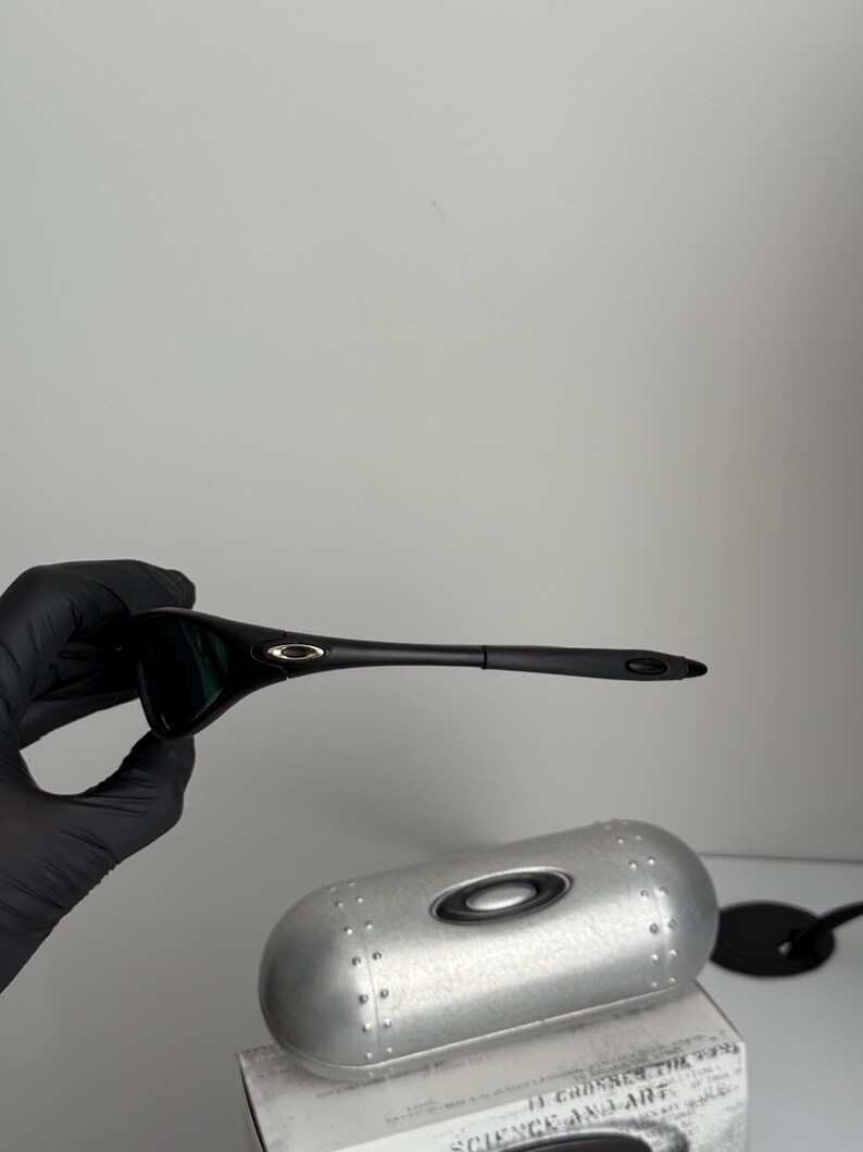 May include: Black sunglasses with a sleek, modern design. The sunglasses have dark lenses and a black frame. A silver case and a black accessory are also visible.