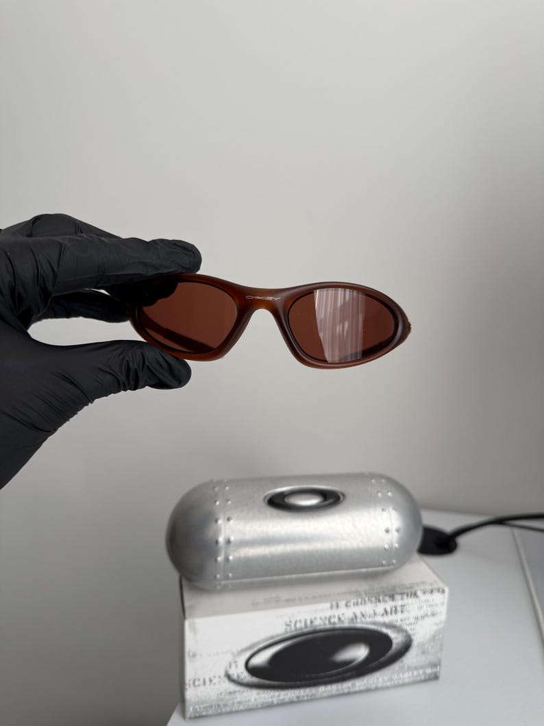 May include: Brown sunglasses with a matching case and box. The sunglasses have a curved frame and lenses. The case is silver and cylindrical. The box has the text "SCIENCE AND ART" and a logo.