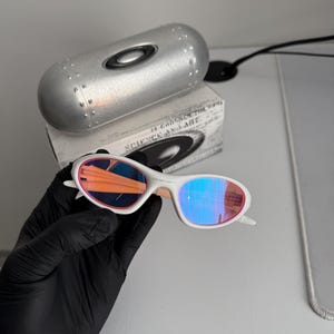 May include: White-framed sunglasses with iridescent lenses, held by a gloved hand. The lenses reflect orange and blue hues. A silver case and a box with the text "SCIENCE AND ART" are in the background.