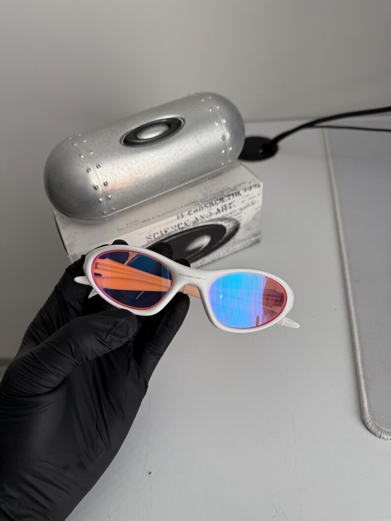 May include: White-framed sunglasses with iridescent lenses, held by a gloved hand. The lenses reflect orange and blue hues. A silver case and a box with the text "SCIENCE AND ART" are in the background.