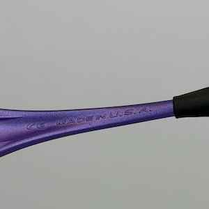 May include: Close-up of a purple eyeglass frame arm with the text "MADE IN U.S.A." and "CE" printed on it. The arm has a gradient color and a black tip.